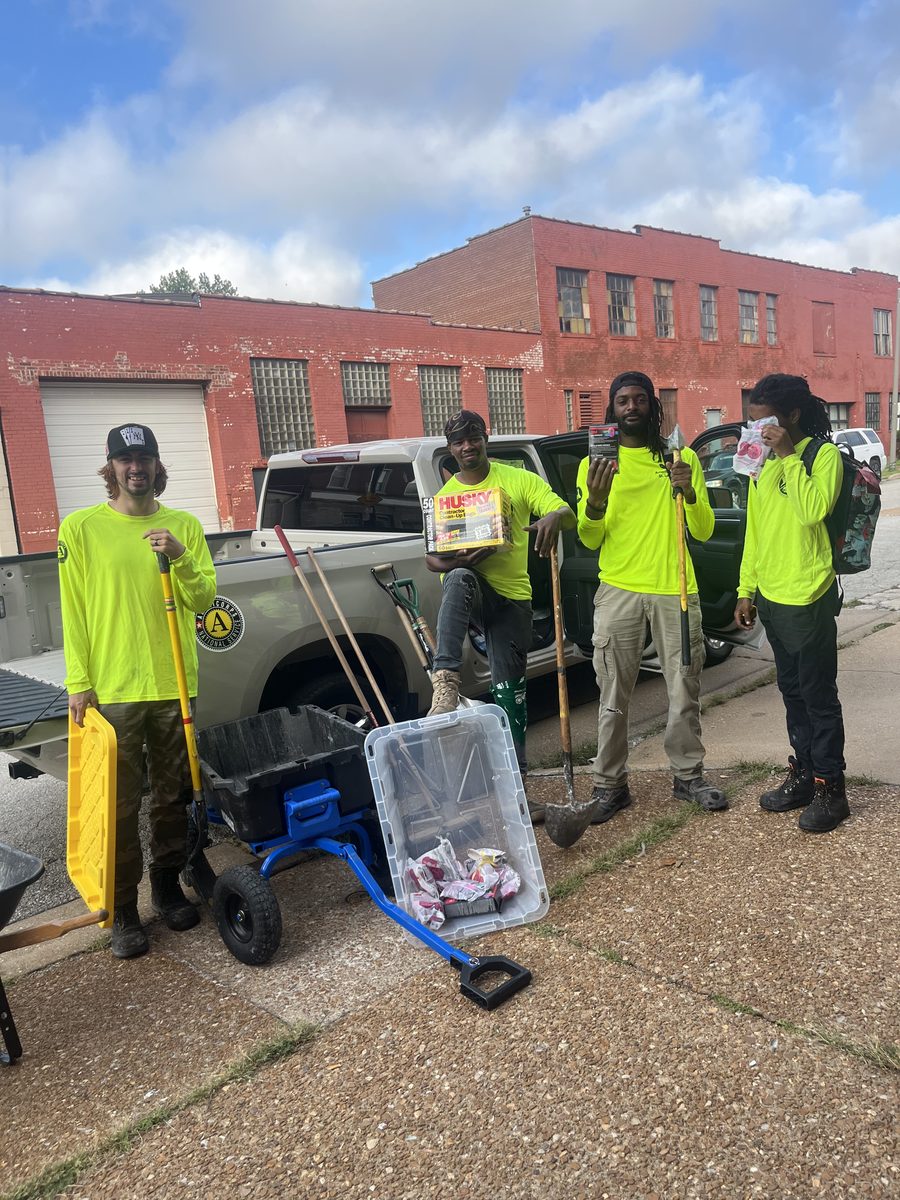 An AmeriCorps crew in hi-vis shirts standing with donated shovels, rakes, and tool kits beside a pickup truck